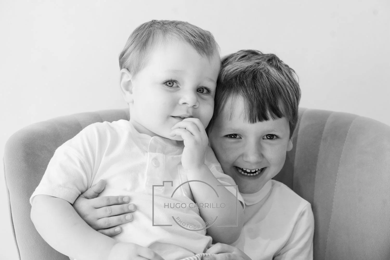 Two young boys sitting close together on a cushioned chair, smiling and embracing each other, in black and white.