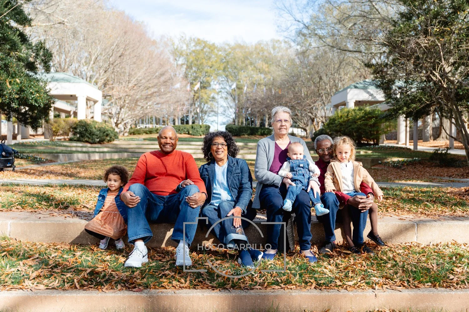 Group of diverse people sitting on a bench outdoors with autumn leaves on the ground and trees in the background.