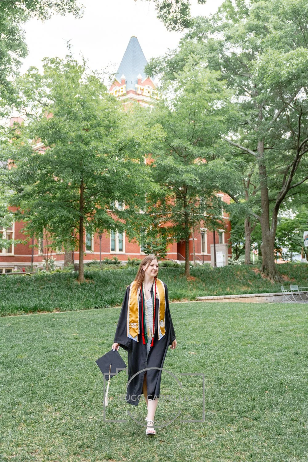 A young woman in a graduation gown and cap, holding her cap in her right hand, walking on a grassy area with trees and a red brick, castle-like building in the background.