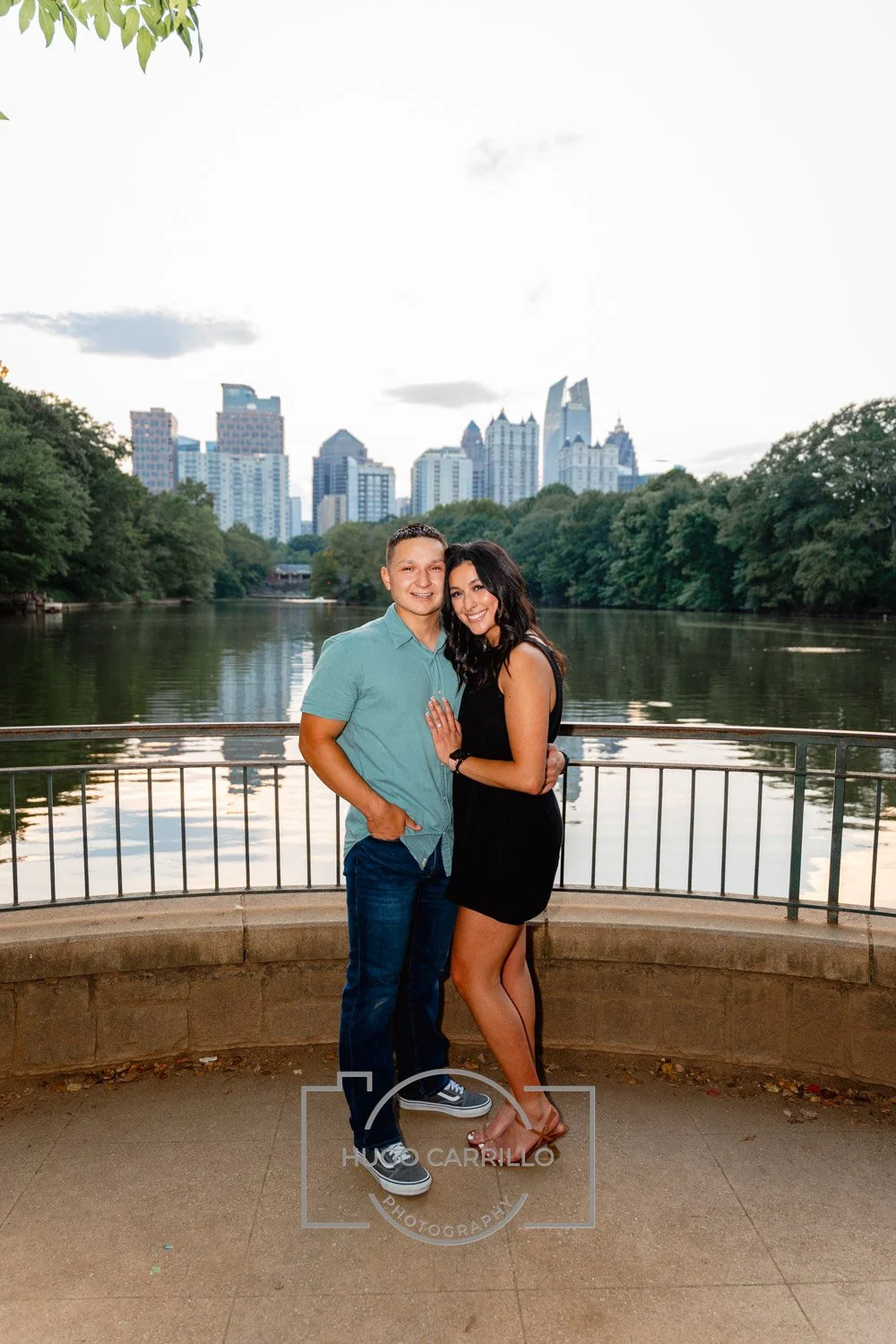 A couple standing close together on a walkway near a river, with a city skyline and green trees in the background. The man is wearing a light blue shirt and jeans, while the woman is dressed in a black dress and heels, smiling at the camera.