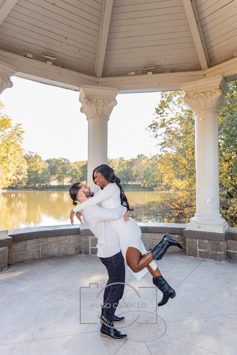 A couple is embracing outdoors in a gazebo by a lake, with the woman wearing a white dress and black boots, and the man in dark pants and a white shirt, during autumn.