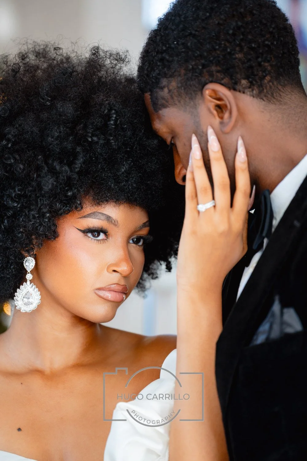 A woman with curly black hair and large earrings gazes at the camera while a man with short curly hair gently touches her face; she is wearing a white dress and the man is dressed in a tuxedo.