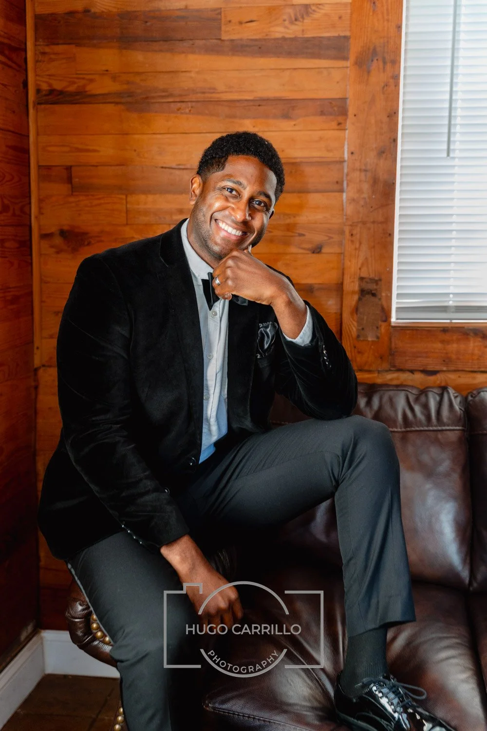A smiling man sitting on a leather sofa in front of a wooden wall, wearing a black blazer, gray pants, a light shirt, and black shoes, with one hand resting on his chin.