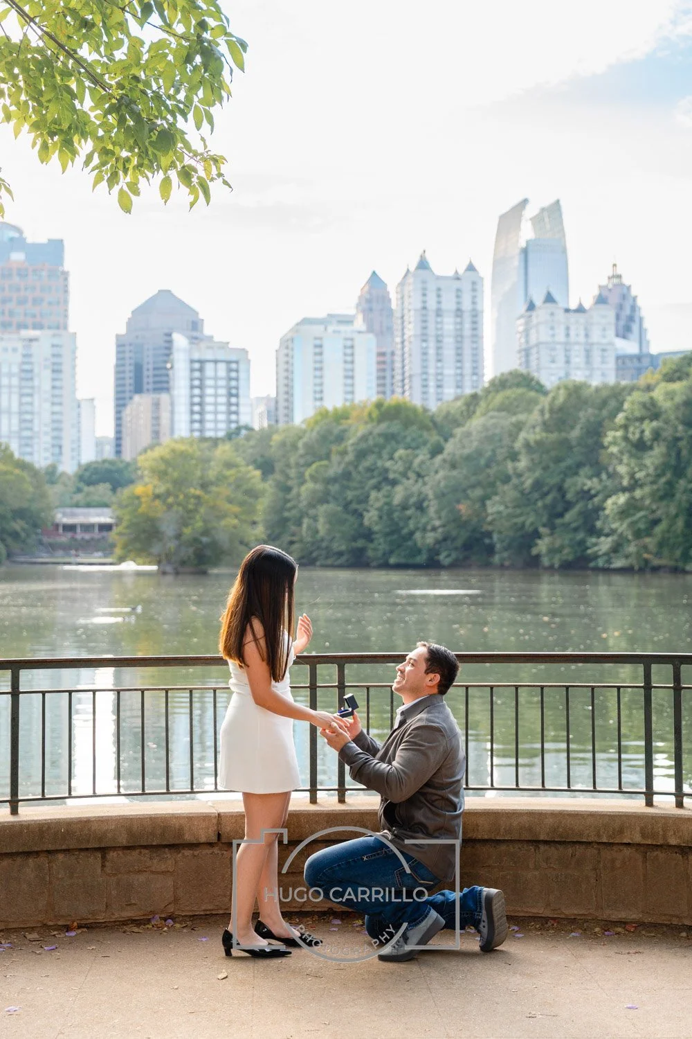 A man proposes marriage to a woman by a river in a park with city buildings in the background.