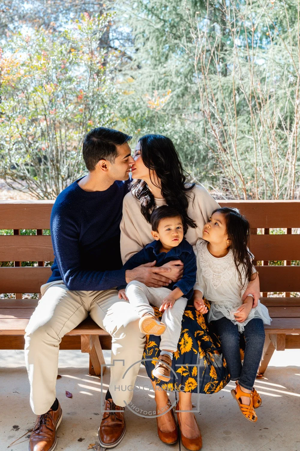 A family of five sitting on a wooden bench outdoors, sharing a moment of affection and love, with a background of trees and bright natural light.