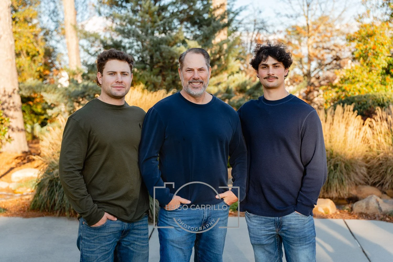 Three men standing outdoors in front of trees with autumn foliage, posing for a photo.