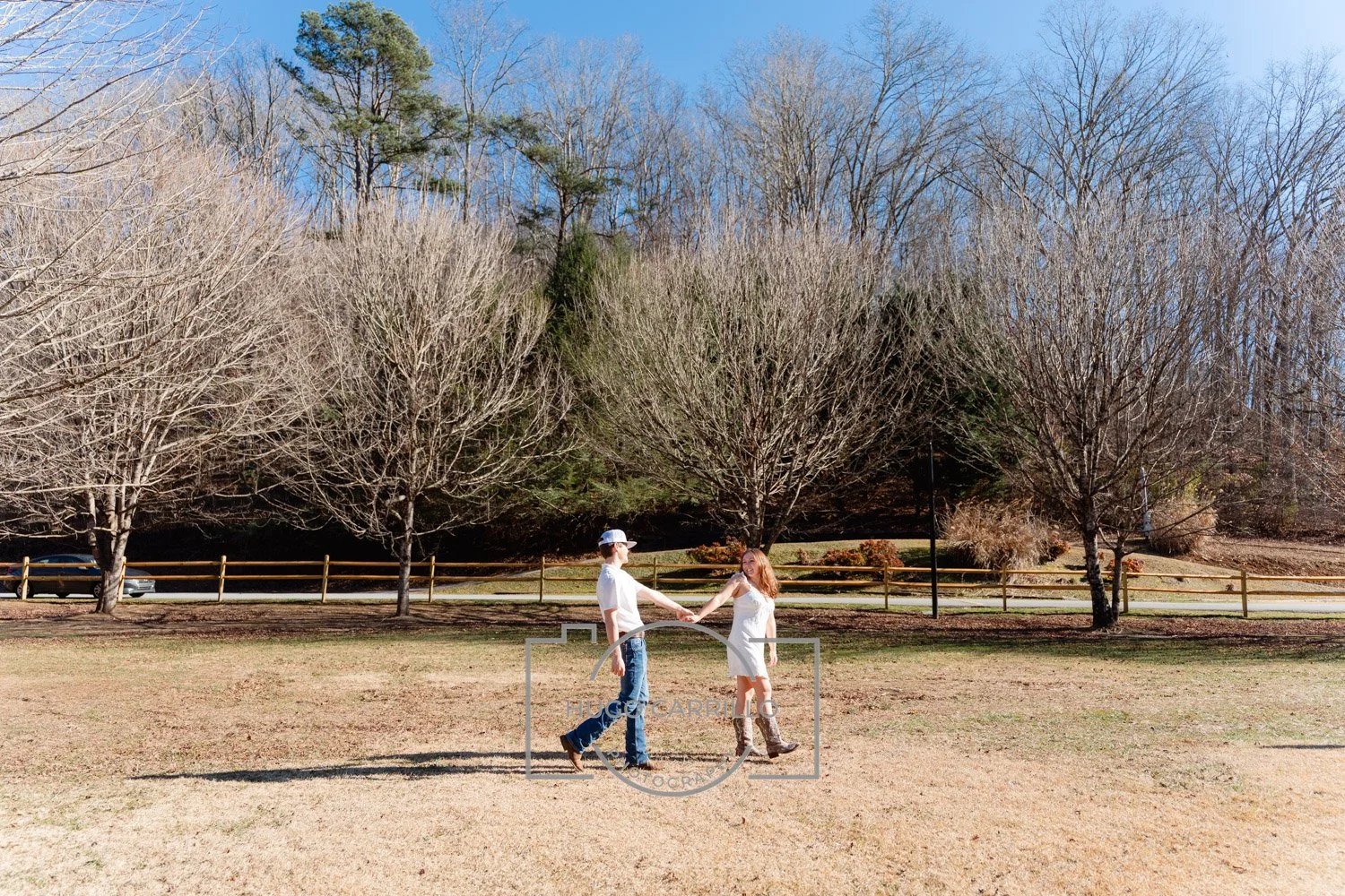 A young man and woman holding hands and walking on a grassy field on a sunny day. The man wears a white t-shirt, jeans, and a baseball cap; the woman wears a white dress and cowboy boots. There are leafless trees and a wooden fence in the background,