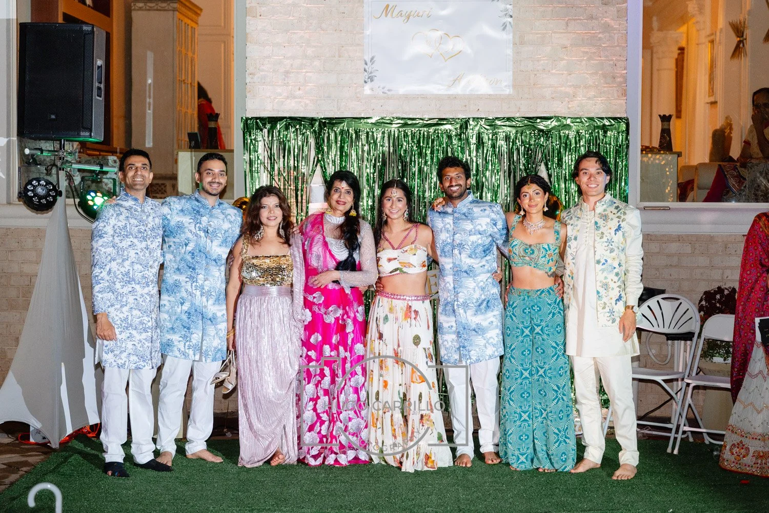 A group of ten people posing together at a celebration, dressed in traditional Indian attire, standing in front of a green shiny backdrop with a white wall and brick wall background.