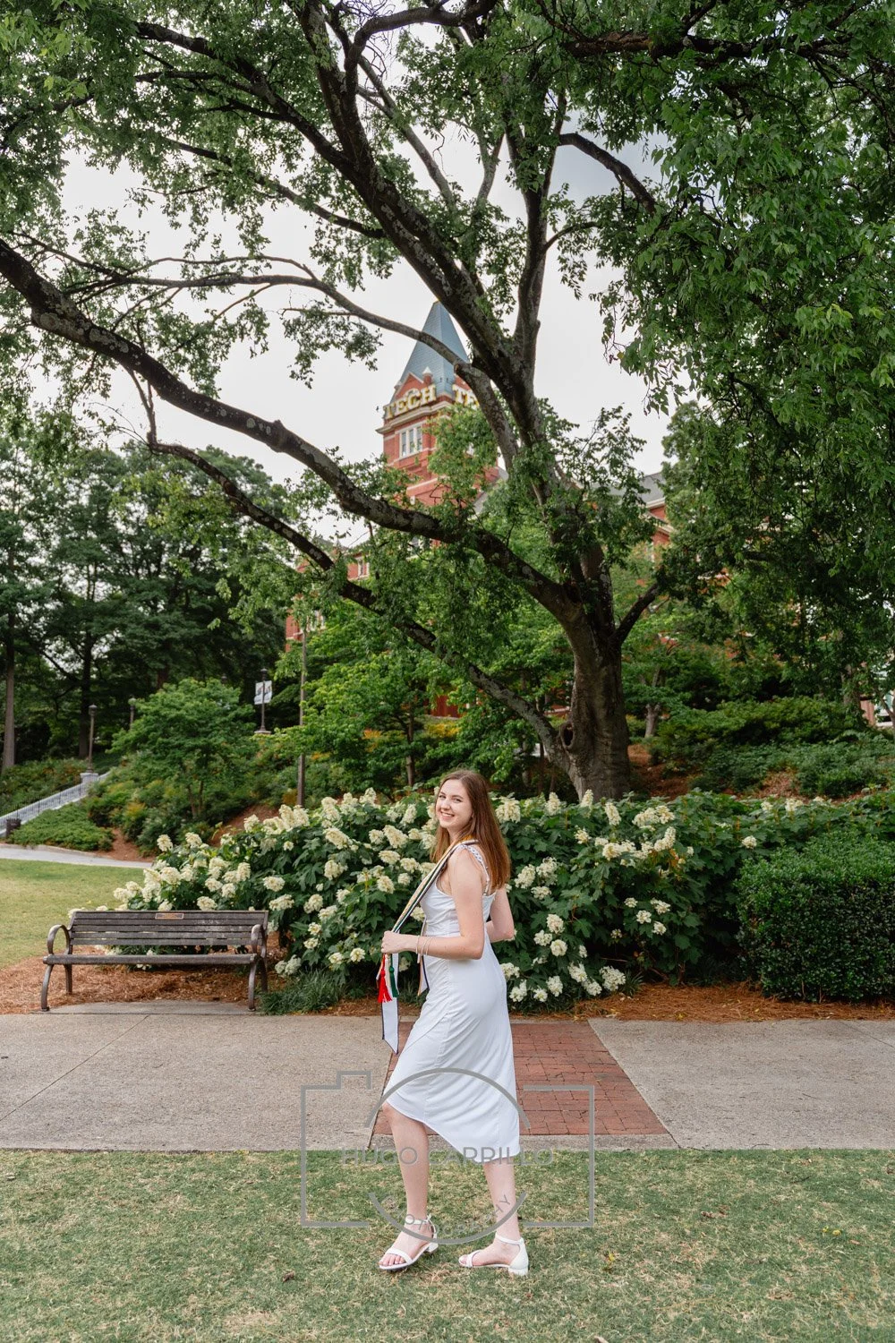 A young woman in a white dress and high heels standing on a sidewalk in a park, smiling at the camera, with a large tree and blooming bushes in the background and a building with a clock tower in the distance.