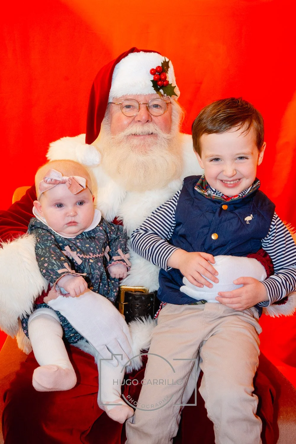 Santa Claus with a white beard and glasses sitting with two children against a red background