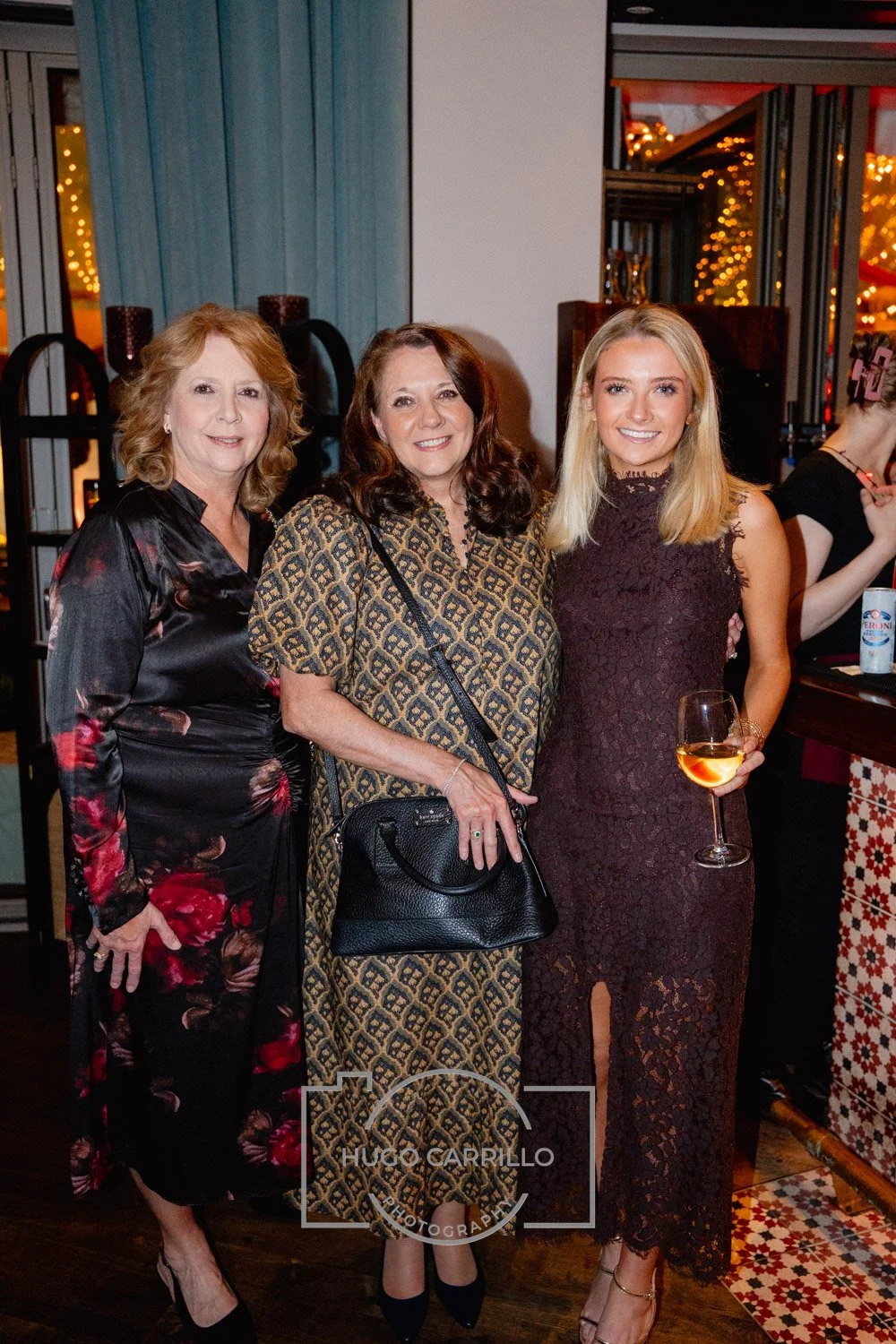 Three women standing together at an indoor event, with two of them holding drinks. The woman on the left wears a dark floral dress, the woman in the middle wears a geometric patterned dress and carries a black purse, and the woman on the right wears 