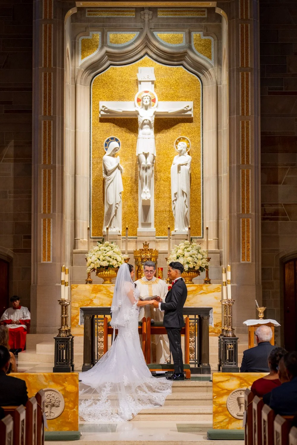 A wedding ceremony taking place inside a church, with a bride and groom standing in front of an altar with a large crucifix and statues of saints. The bride wears a white wedding gown and veil, and the groom wears a black tuxedo. An officiant is standing between them, holding a book, while an altar boy sits to the side.
