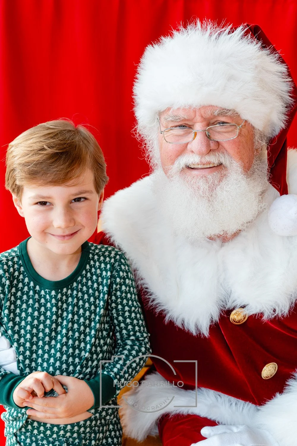 A young boy smiling next to Santa Claus, who is dressed in a red velvet suit with white fur trim, glasses, and a Santa hat, in front of a red curtain.