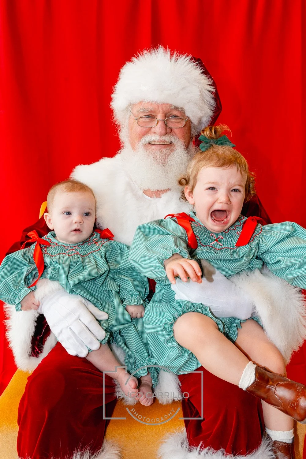 Santa Claus with two young children, one crying and one looking surprised, against a red backdrop.
