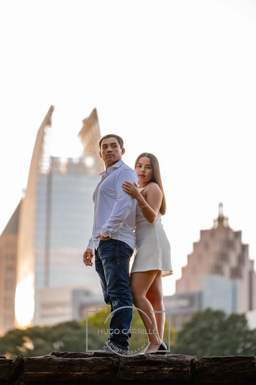A young man and woman standing close together on a rocky surface in an urban park with city skyscrapers in the background during sunset.