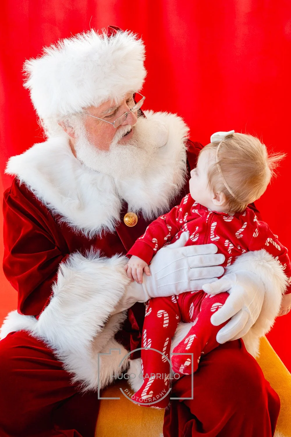 Santa Claus holding a young girl with blonde hair and a bow, both dressed in Christmas pajamas, against a red background.