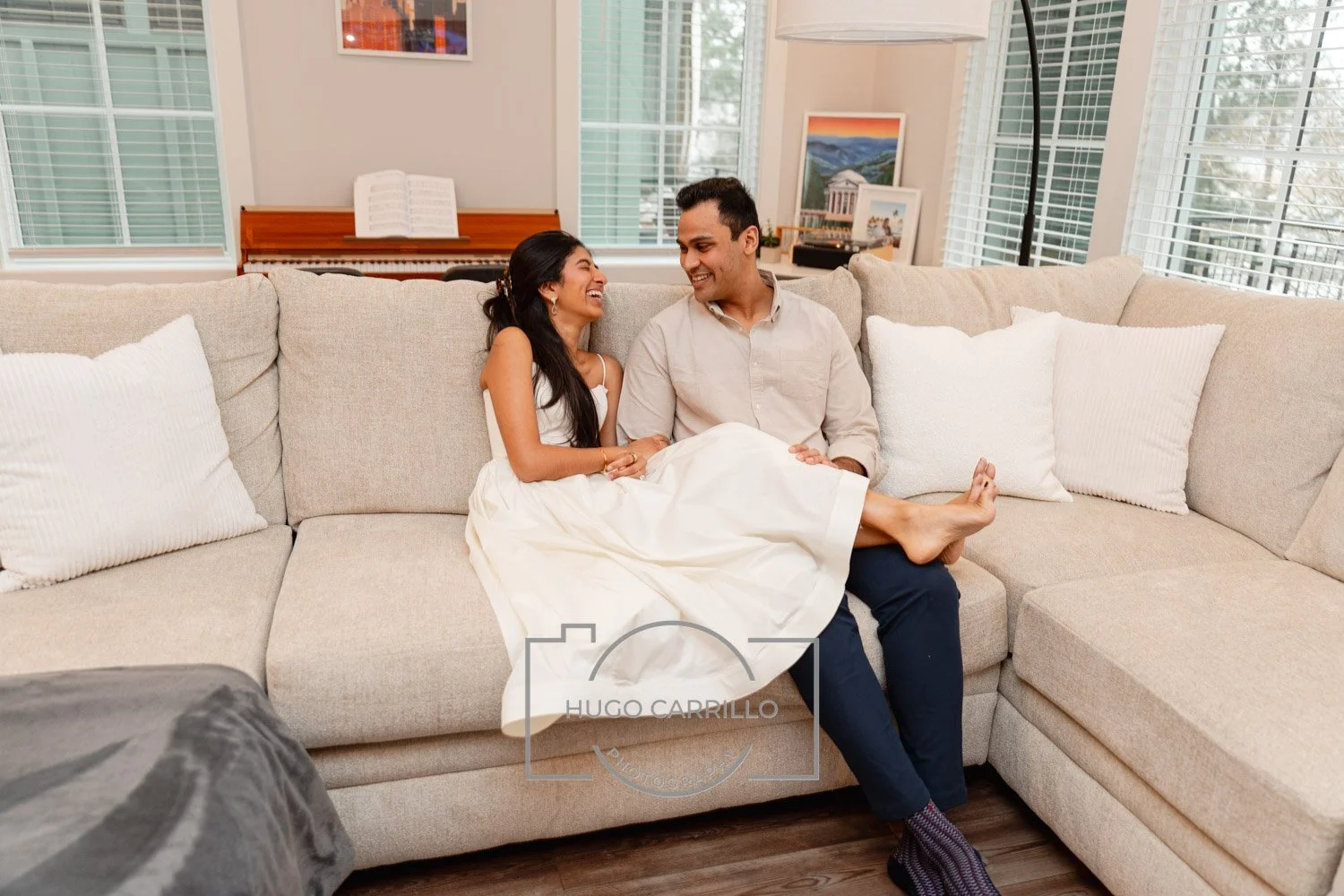 A couple sitting on a beige sectional sofa in a living room, smiling and looking at each other, with the woman sitting on the man's lap and both appearing happy.
