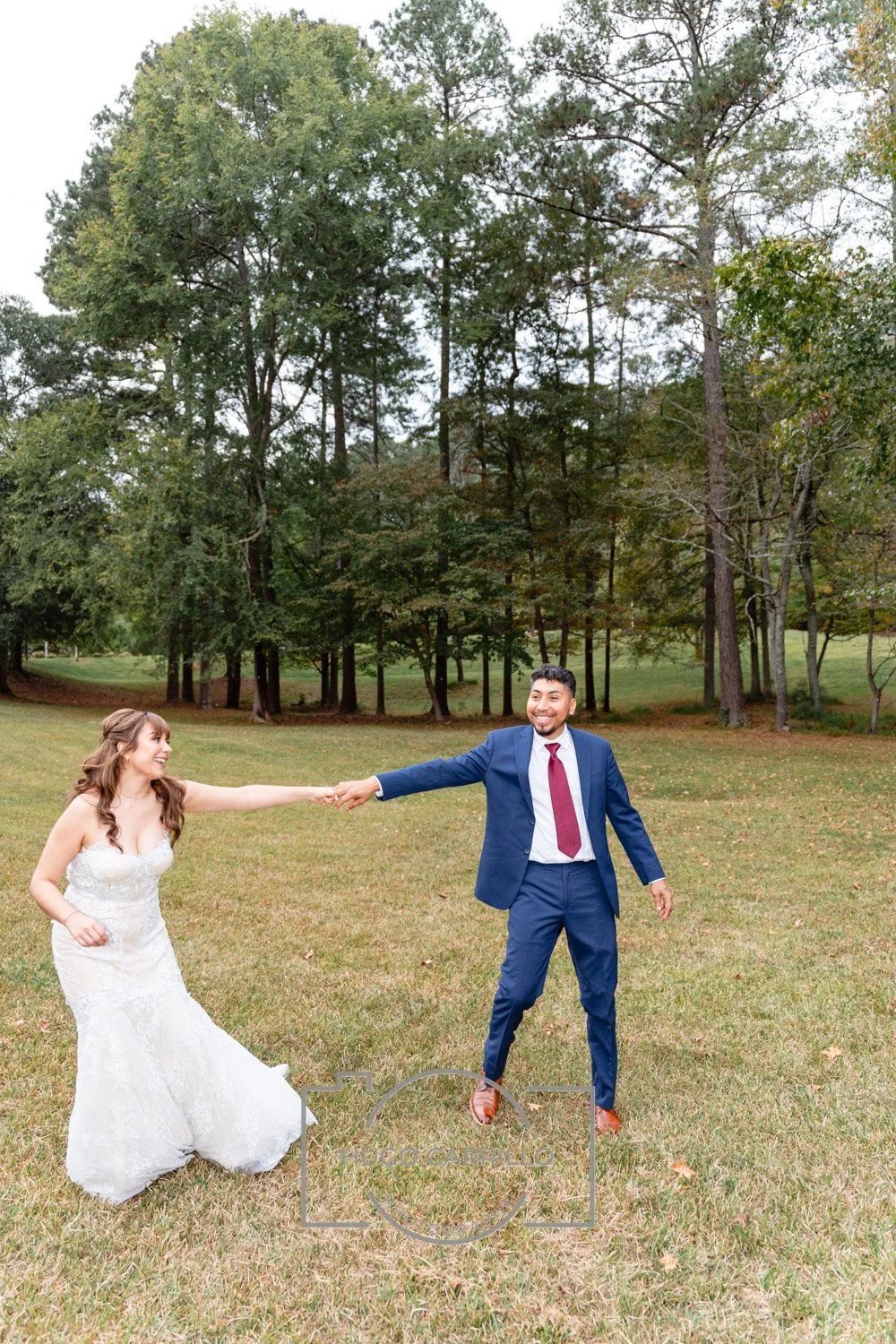 A bride in a white wedding gown and a groom in a blue suit are holding hands and smiling in a grassy park with trees in the background.