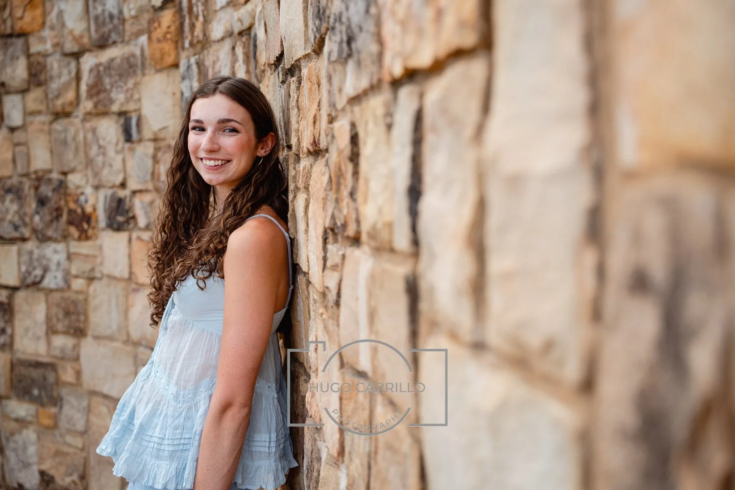 A young woman with long, curly brown hair smiling and leaning against a textured stone wall.