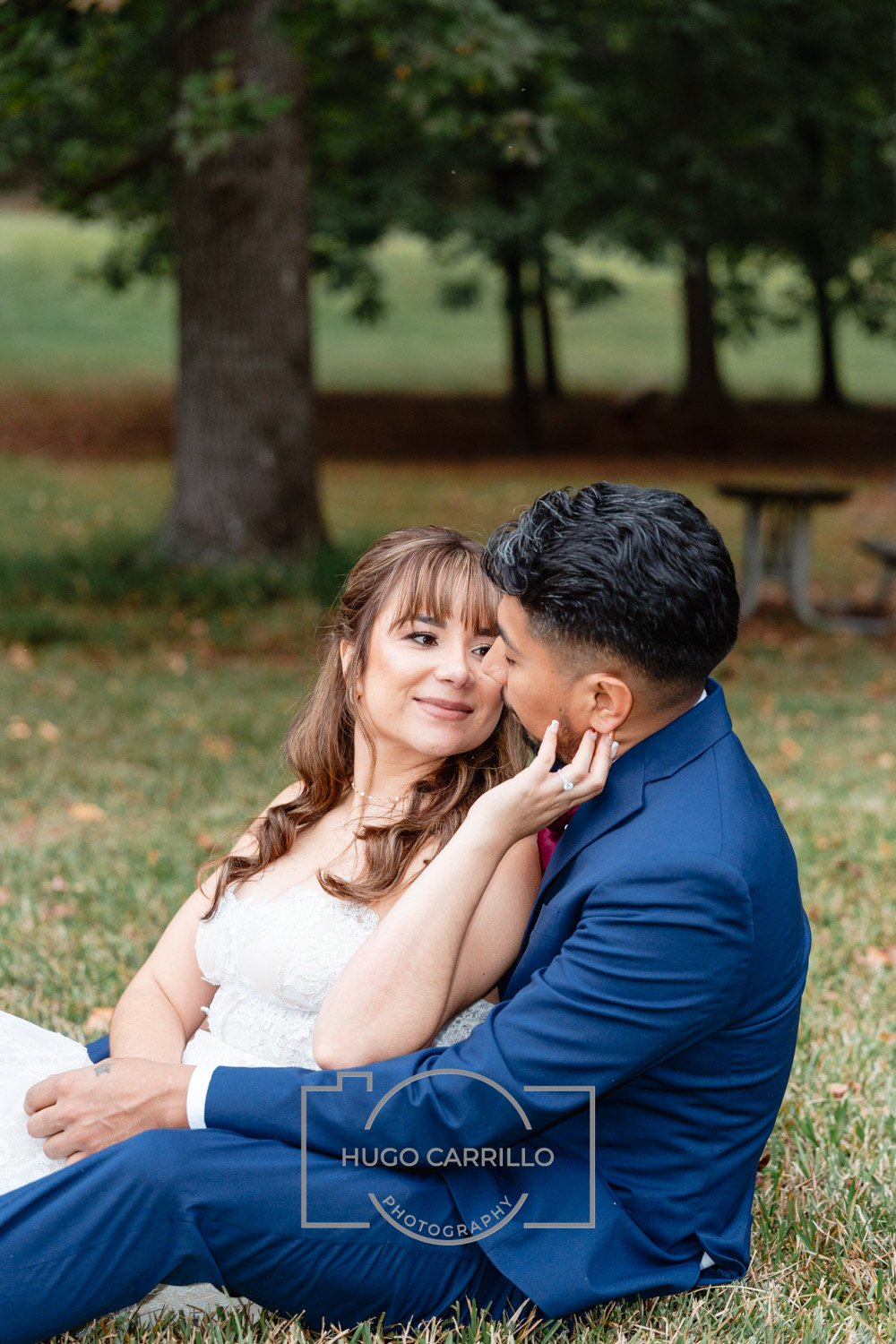 A couple dressed in wedding attire sitting on the grass, with the woman gently touching the man's face, in a park with trees in the background.