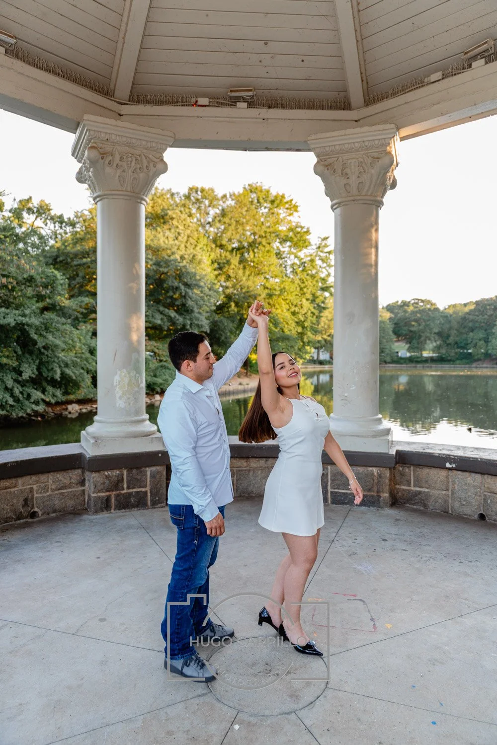A couple dancing under a gazebo near a lake, with trees in the background and the woman smiling.