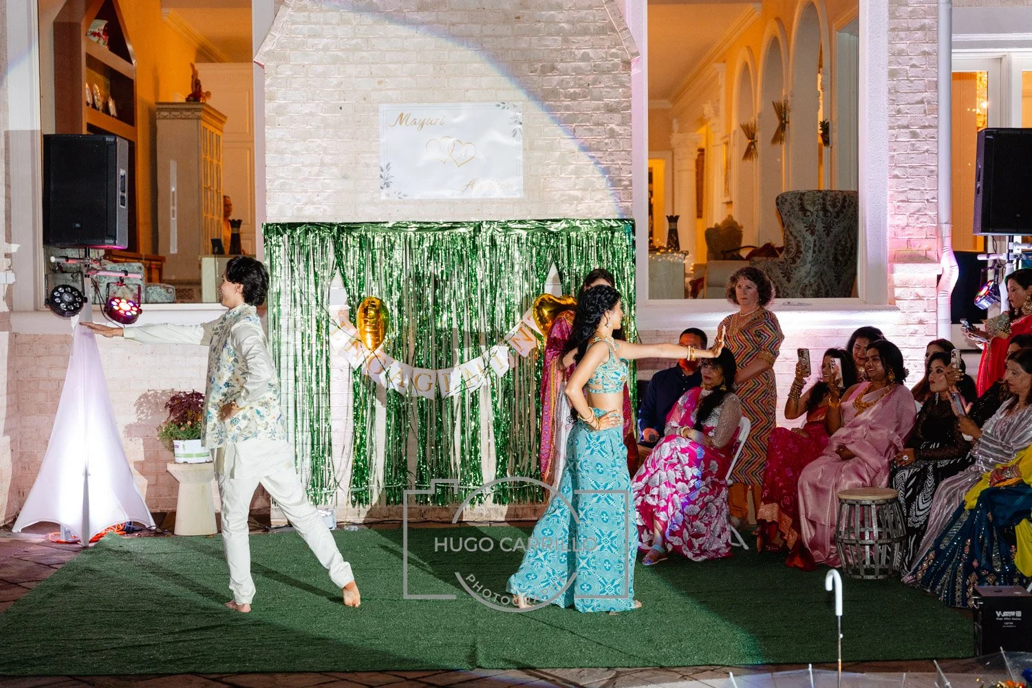 A cultural dance performance at a celebration event in an indoor/outdoor venue with guests seated and watching. The performers are dressed in traditional Indian attire, with a green backdrop and decorative balloons, and a banner indicating a birthday
