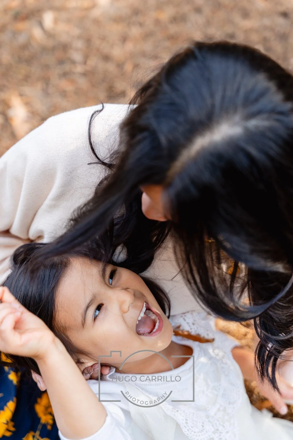 A woman with long black hair leaning over a young girl with black hair, smiling and raising her hand, on a ground covered with fallen leaves.