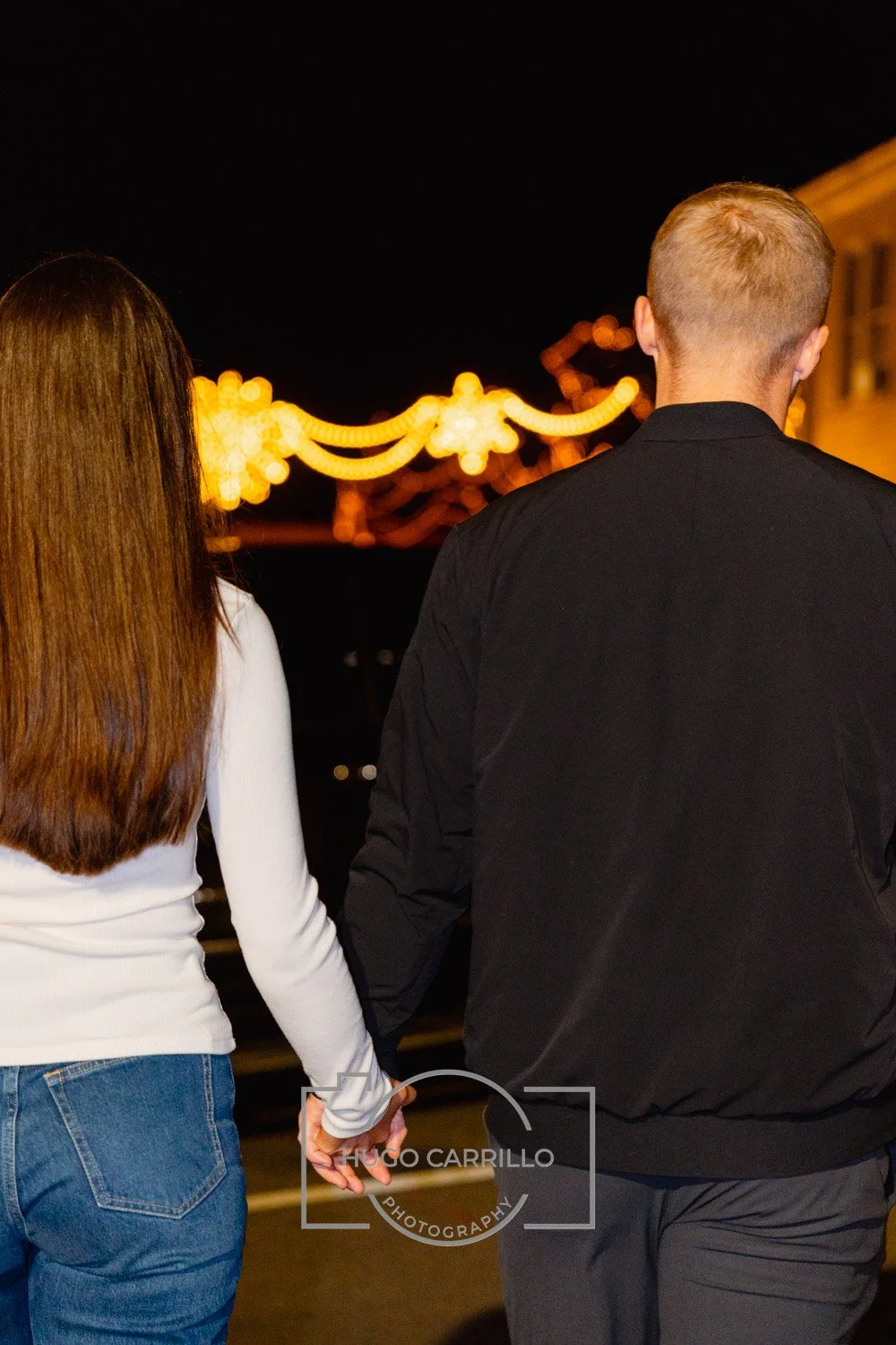 A couple holding hands at night, facing away from the camera, with festive string lights glowing in the background.