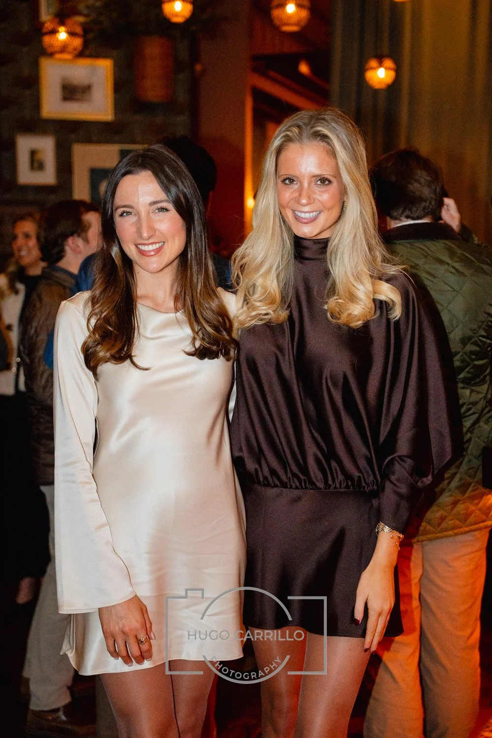 Two women in stylish dresses standing close together, smiling at the camera during an indoor event with warm lighting and decorative lanterns.