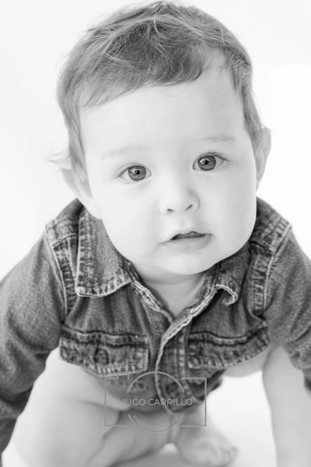 Black and white photo of a baby with big eyes, short hair, and wearing a denim jacket, looking directly at the camera.