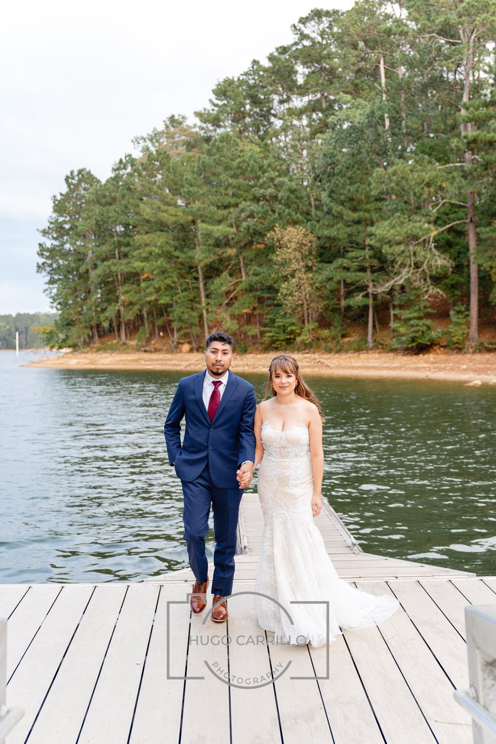 Bride and groom walking hand in hand on a dock by a lake surrounded by green trees.