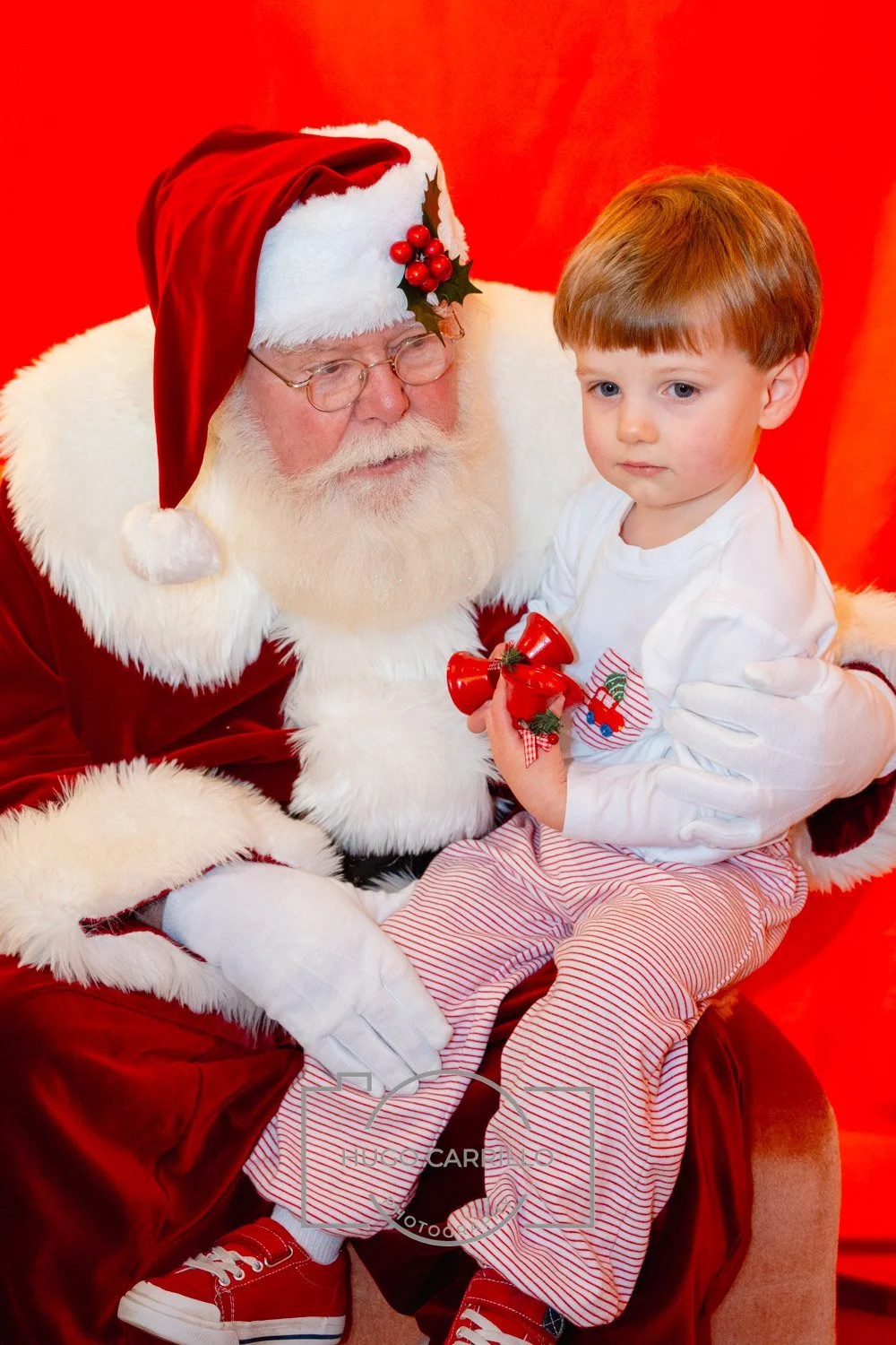 A young boy sitting on Santa Claus's lap during a Christmas event, holding red Christmas bells, with Santa in his traditional red suit, hat, and white gloves, both against a red background.