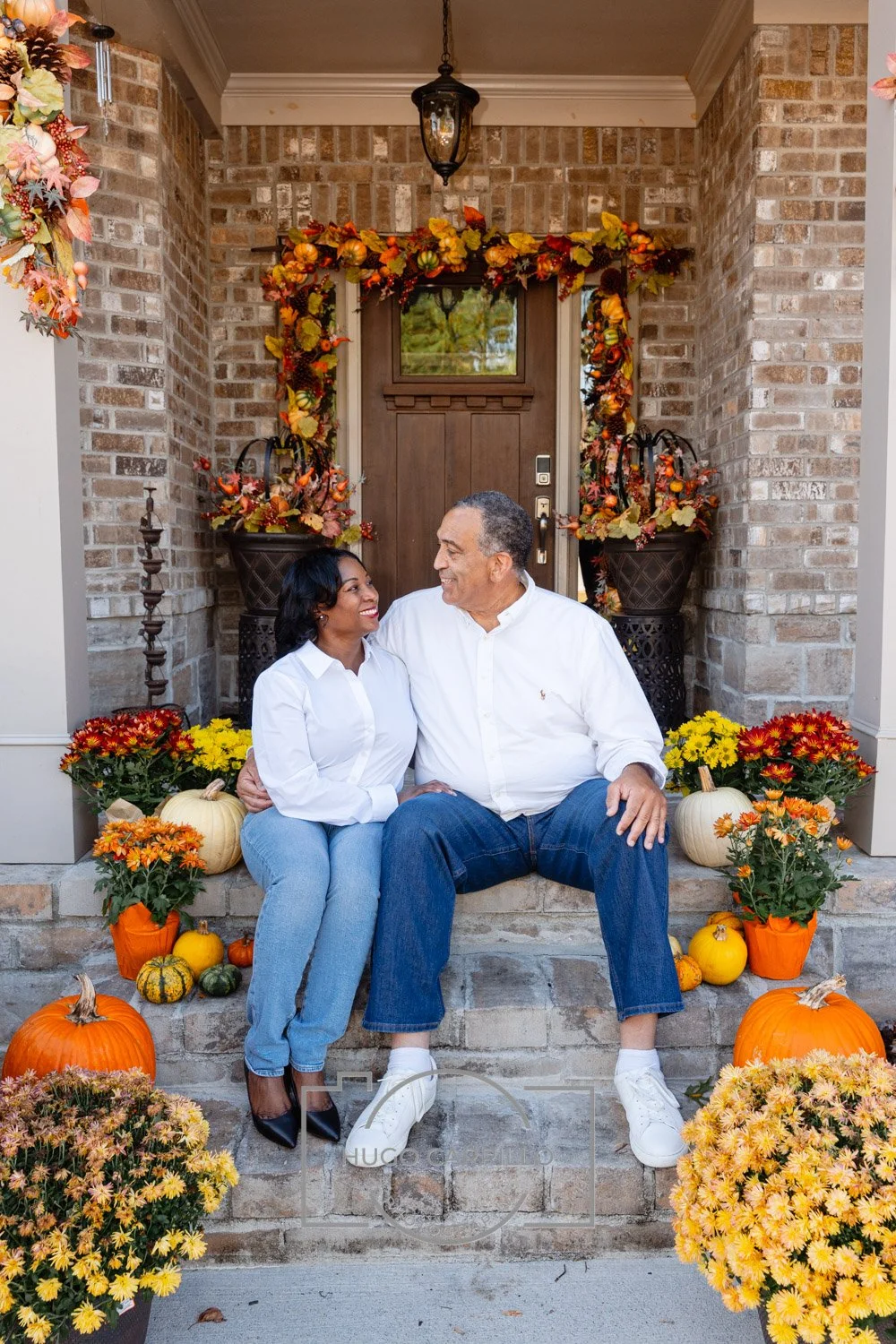 An elderly man and a young woman sitting on the front porch steps of a brick house, smiling at each other surrounded by fall pumpkins and flowers.