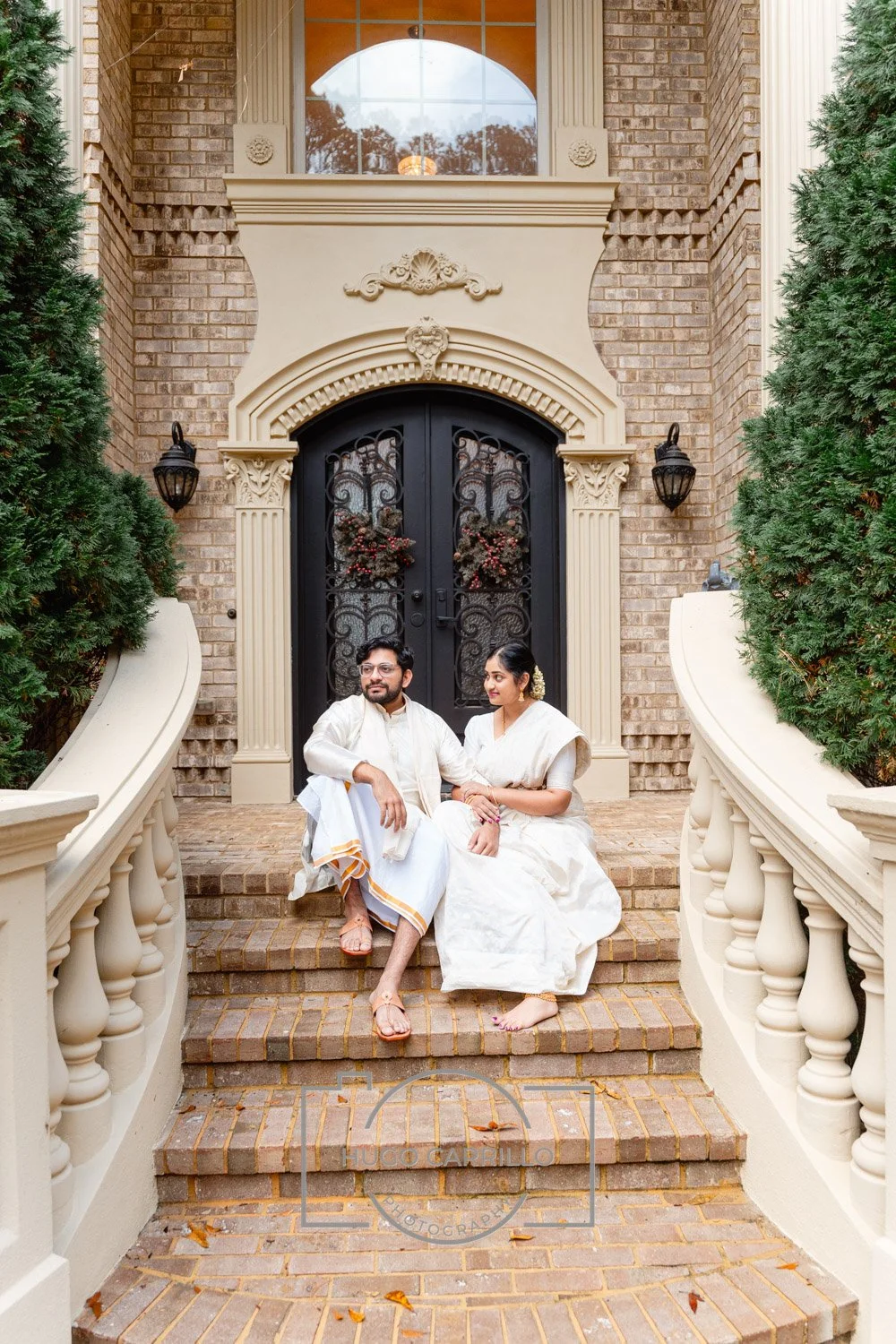 A couple dressed in traditional white Indian attire sitting on brick stairs in front of a large ornate black door with wrought iron design, flanked by greenery and decorative beige architecture.