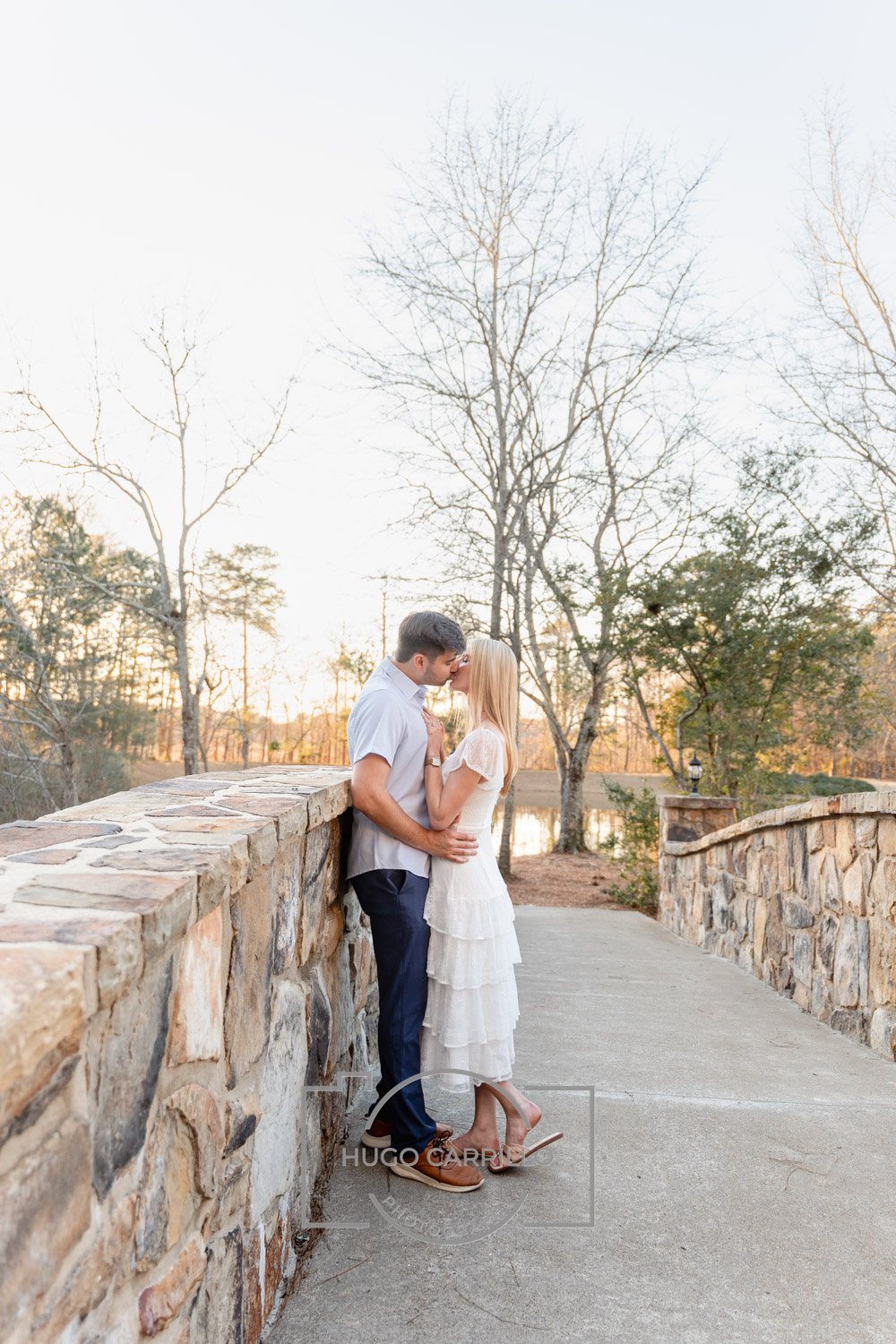 A romantic couple sharing a kiss on a stone bridge during sunset, surrounded by bare trees and a pond in the background.