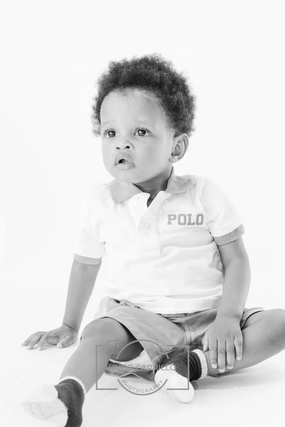 Black and white photo of a young child with curly hair, sitting on the floor, wearing a polo shirt and shorts, looking to the side.