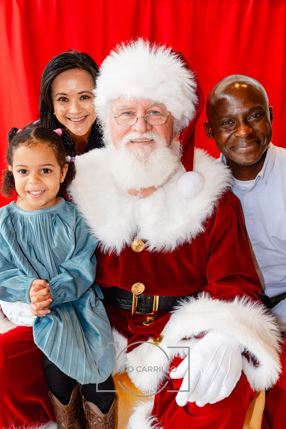 A diverse group of four people, including a young girl, a woman, a man dressed as Santa Claus, and a man, posing together in front of a red curtain, smiling for a holiday photo.