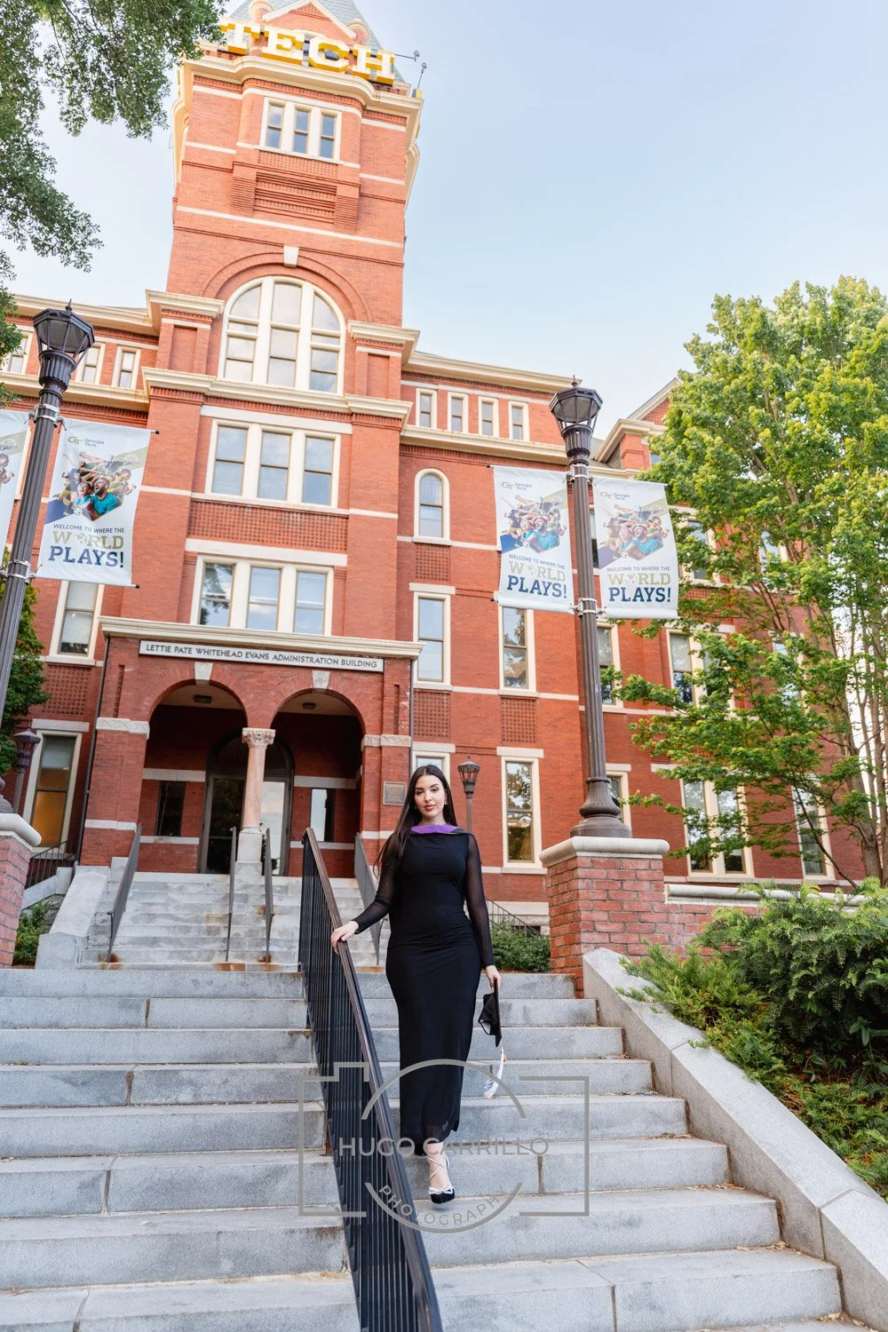 A woman in a long black dress walking down a stone staircase outside a large red brick building with a tower, banners, and trees, during daytime.
