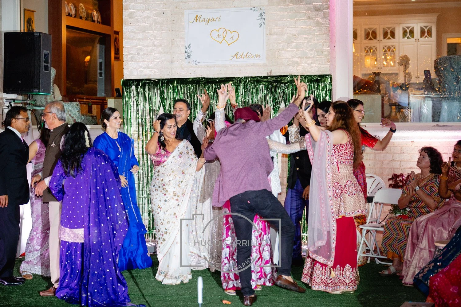 Group of people at a celebration dancing and enjoying, with women in traditional colorful dresses and men in suits, under a decorated backdrop with a sign that reads 'Mayuri and Addison'.