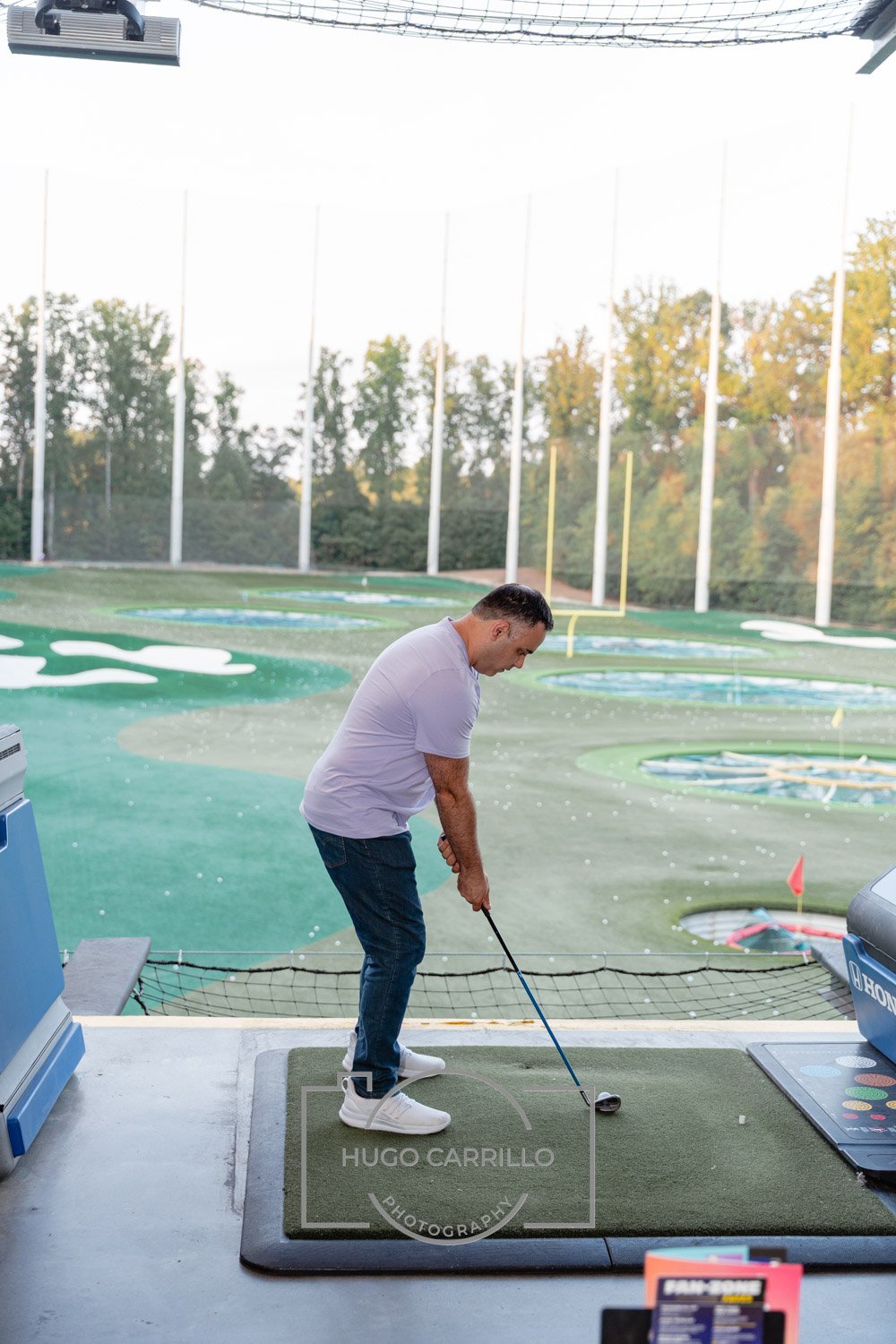Person preparing to hit a golf ball at an indoor driving range with multiple target rings and trees visible outside.