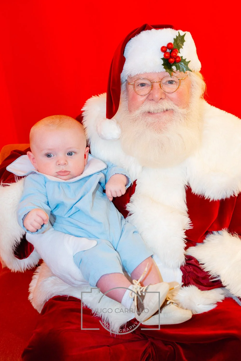 A young baby and a man dressed as Santa Claus posing for a Christmas photo against a red background. Santa is wearing a traditional red suit with white trim and has a sprig of holly on his hat.