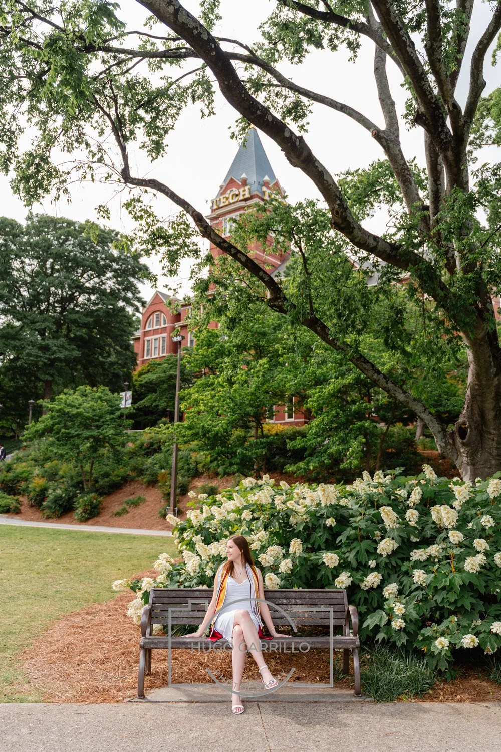 A young woman sits on a park bench beside a large bush with white flowers, in front of an old brick building with a tower, partially obscured by trees, on a bright day.