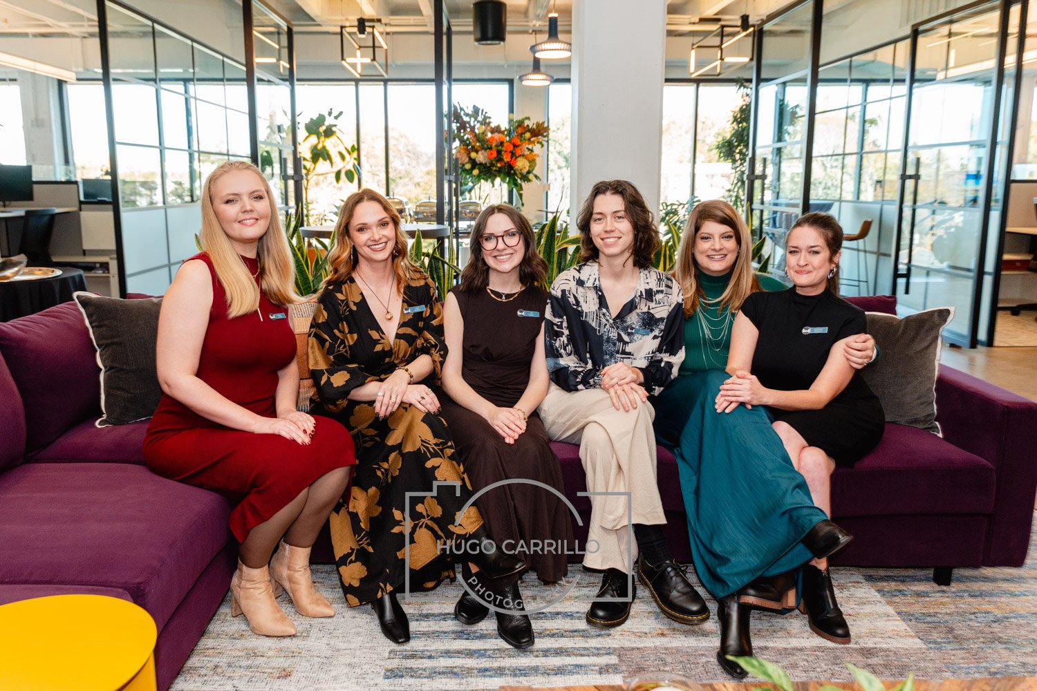 Group of six women sitting on a purple sofa in an office or lounge setting, smiling and looking at the camera, with plants and large windows in the background.