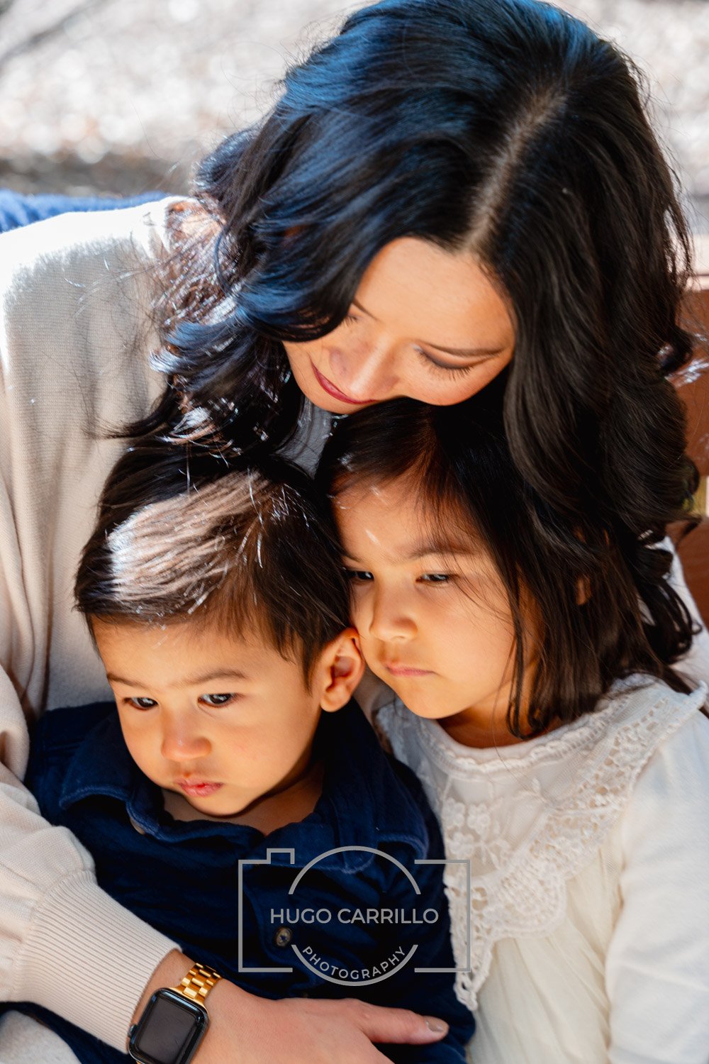 A woman with black wavy hair embracing two young children, a boy and a girl, in a close, tender moment.