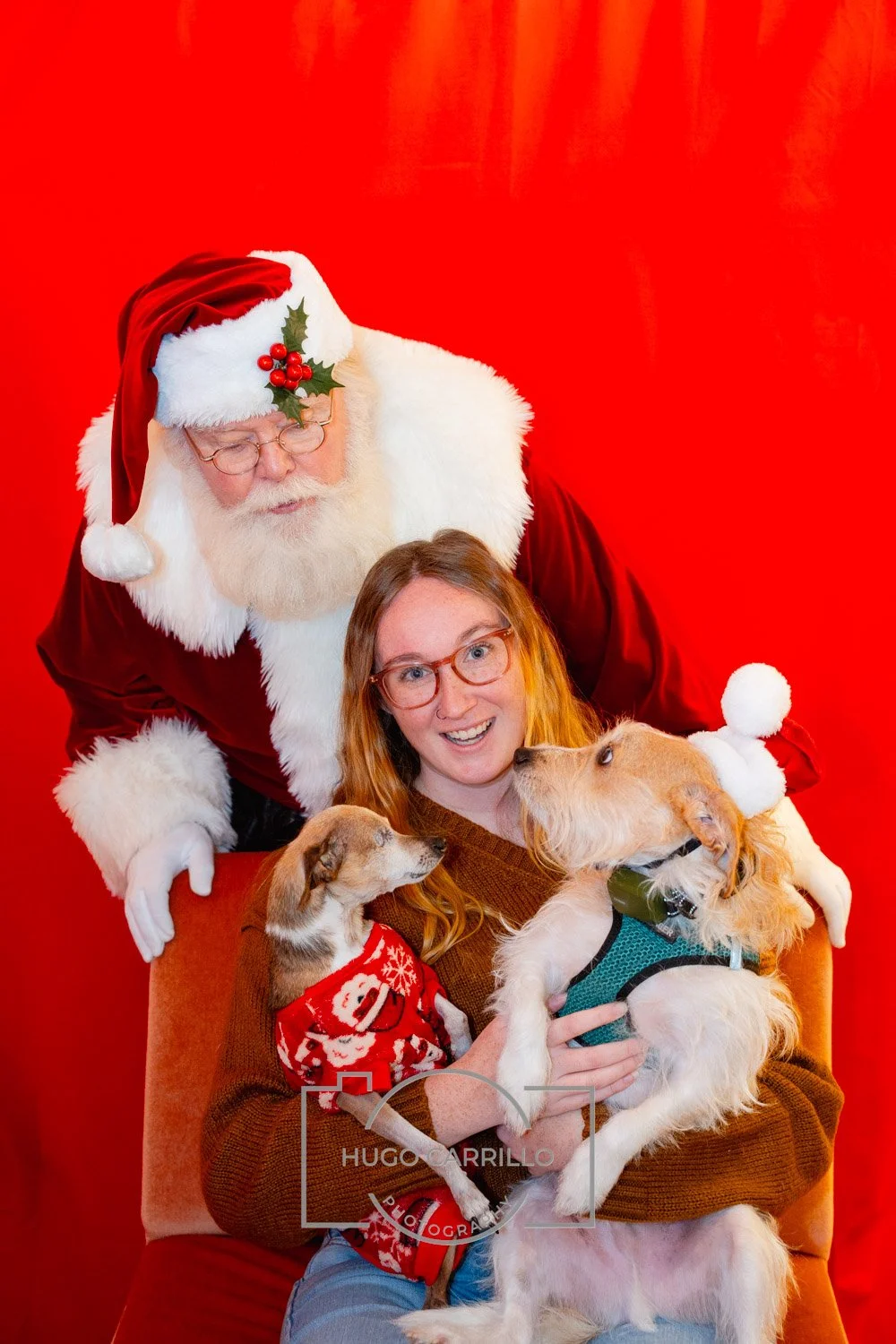 A woman with red hair and glasses sitting on a chair with two dogs, one wearing a Christmas sweater. Santa Claus stands behind her, leaning over, with a festive holiday background.