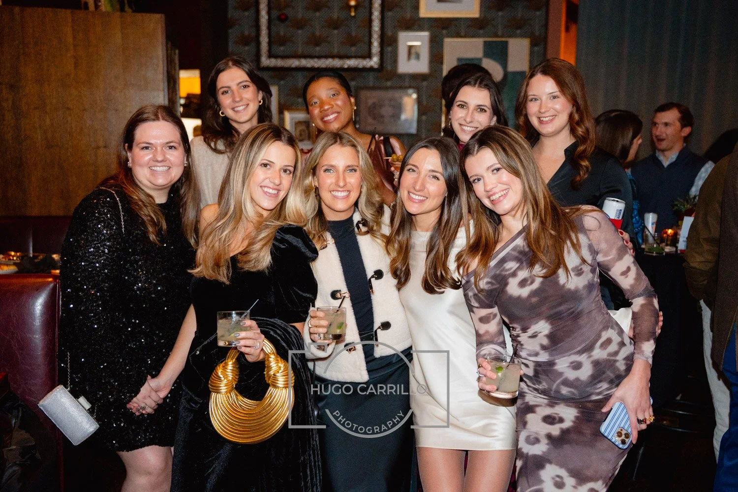 Group of women at a celebration or party, smiling and holding drinks in a social setting with dim lighting and artwork on the wall.