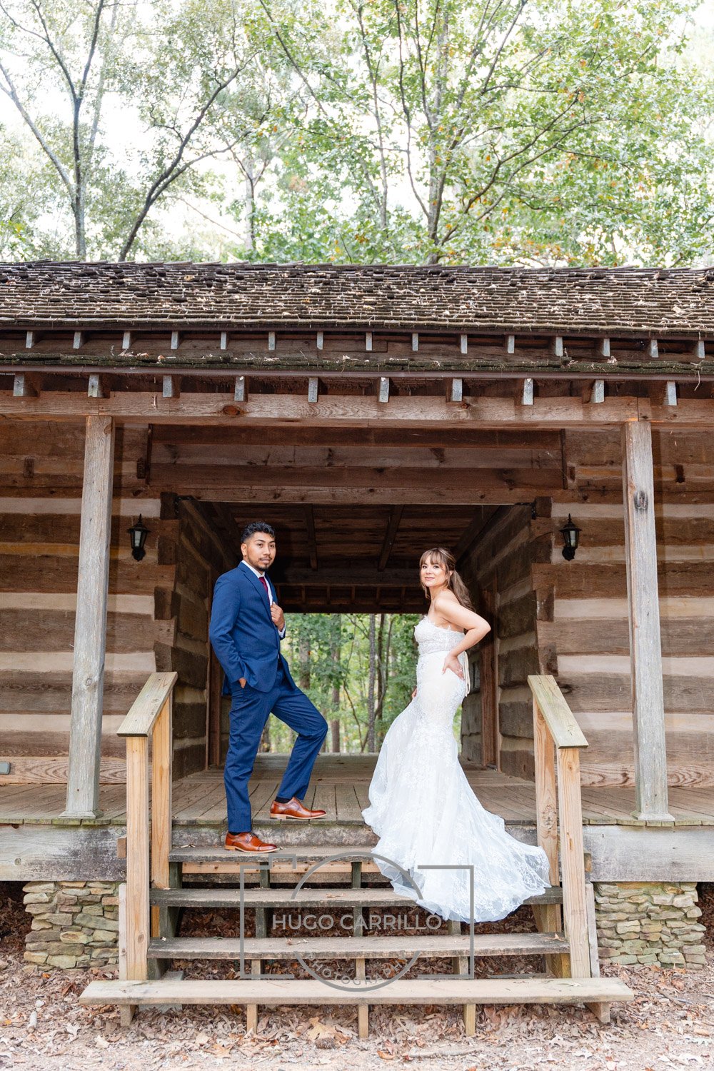 A bride in a white wedding dress and a groom in a blue suit stand on the steps of a rustic wooden building in an outdoor setting with trees in the background.
