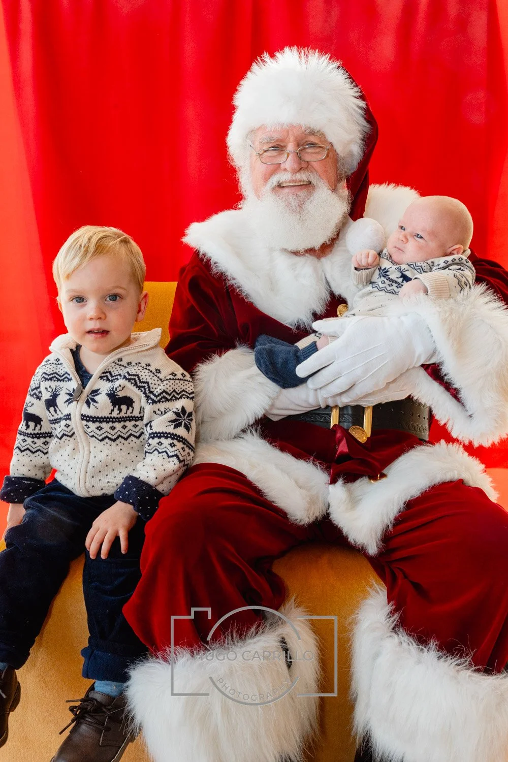 Santa Claus holding a baby and sitting next to a young boy, with a red backdrop.