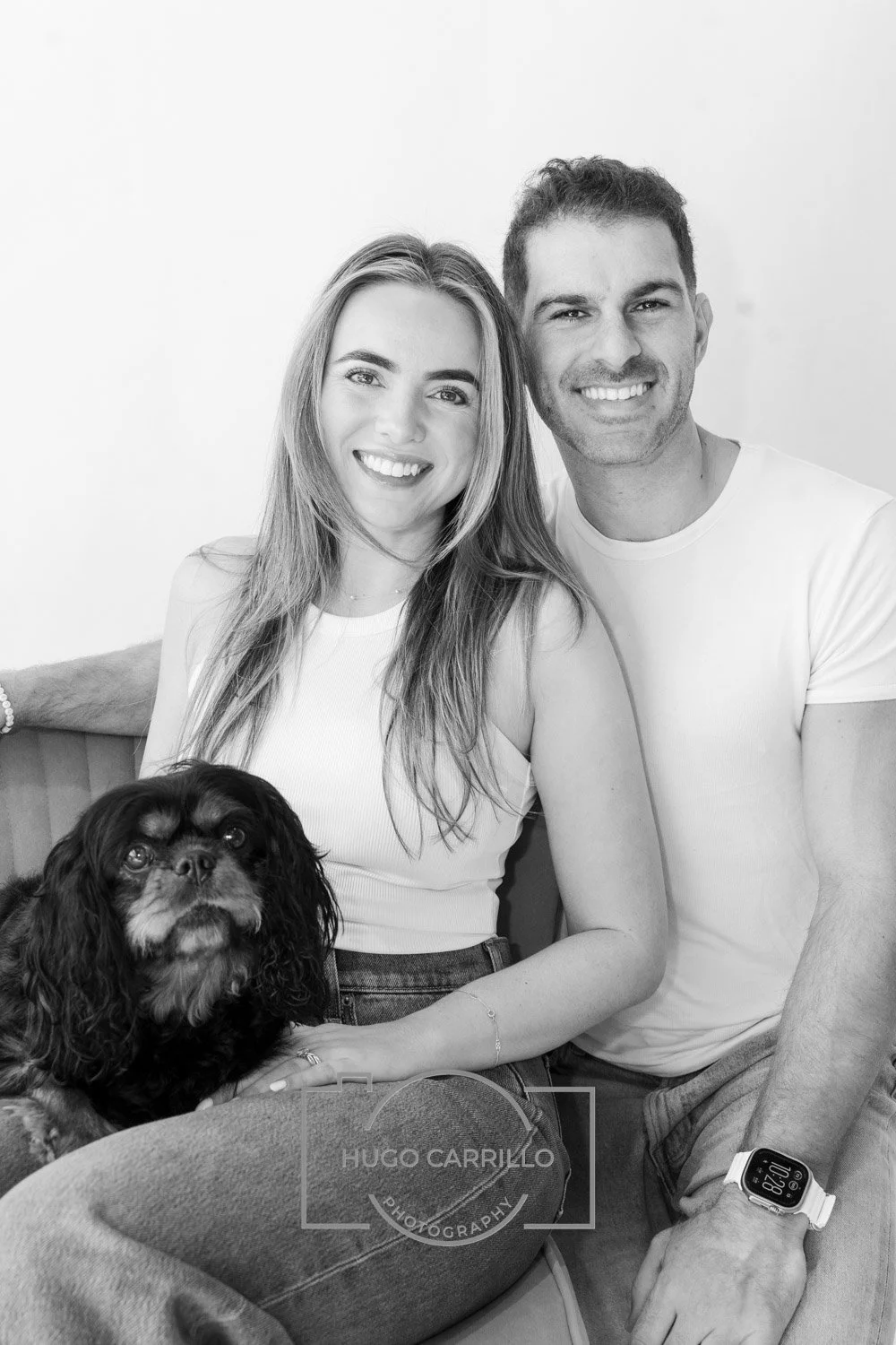 A smiling young woman with long hair and a man with short hair sitting on a couch with a small, dark-colored dog with long ears. The woman is wearing a sleeveless white top, and the man is wearing a white t-shirt and a wristwatch. The photo is in bla