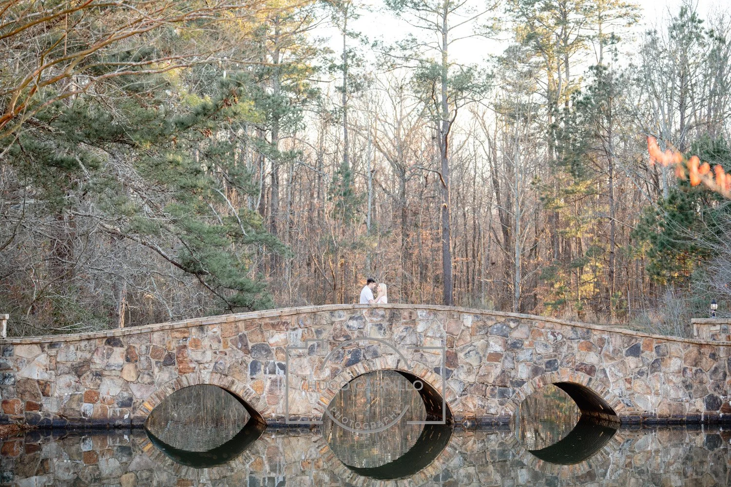 A couple standing on a stone bridge over a river, surrounded by trees with some autumn leaves. The couple appears to be embracing.